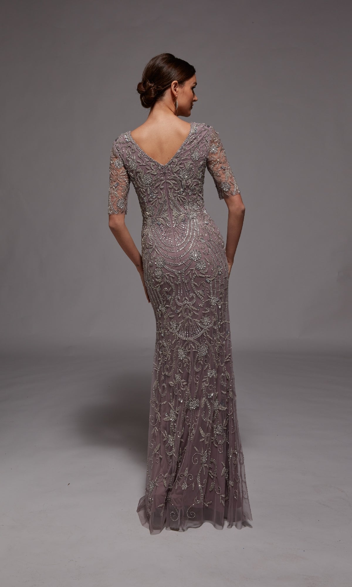 A woman stands with her back to the camera, wearing the Alyce 27824 long formal dress in mauve, featuring floor-length beaded embroidery and lace, elbow-length sleeves, and a V-shaped back—ideal for formal occasions.